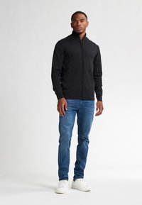 Schwarzer Strickjacke mit Reißverschluss und geripptem Kragen, über Jeans getragen. Das Model steht vor einem schlichten weißen Hintergrund. Weiße Sneakers sind sichtbar.