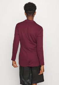 Maroon kavaj med ett texturerat tyg, kolt snitt och långa ärmar; bärs över svarta, löst sittande shorts.