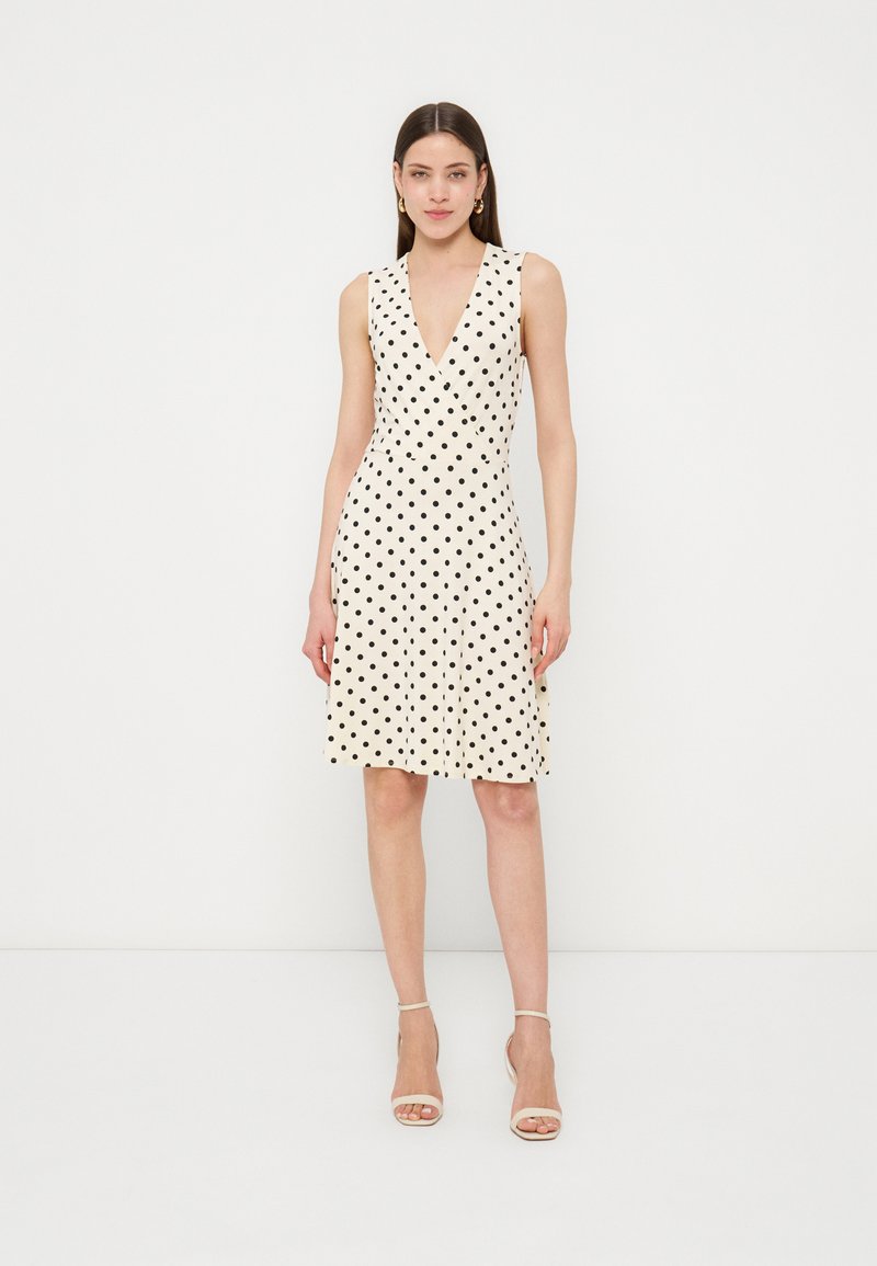 Femme portant une robe blanche sans manches à pois noirs et des sandales beige à talons hauts, debout devant un fond blanc uni.