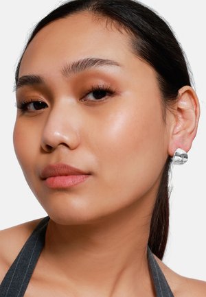 Silberne Ohrringe mit einer glänzenden, glatten Oberfläche, die ein abgerundetes, modernes Design aufweisen. Das Modell hat einen neutralen Gesichtsausdruck und zarte Make-up.