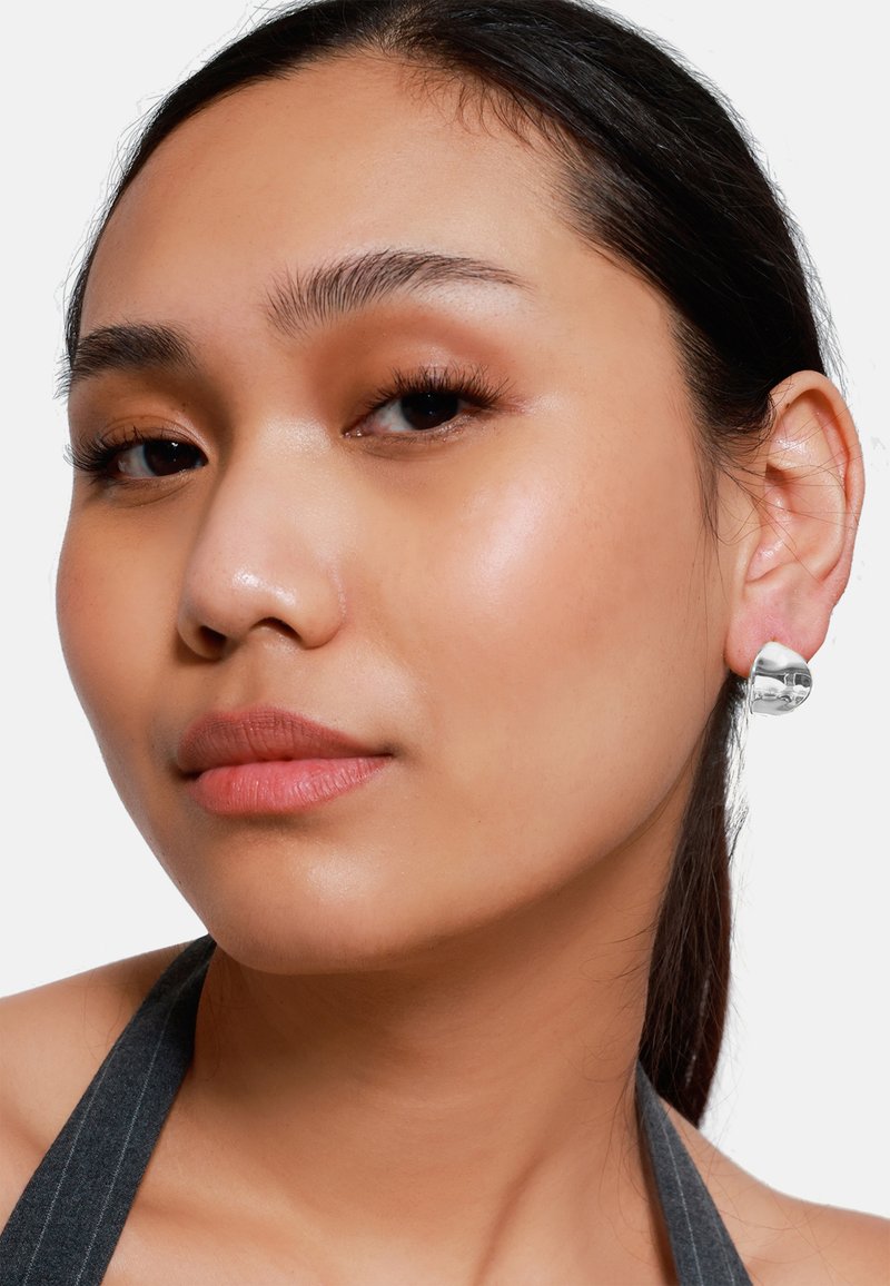 Silberne Ohrringe mit einer glänzenden, glatten Oberfläche, die ein abgerundetes, modernes Design aufweisen. Das Modell hat einen neutralen Gesichtsausdruck und zarte Make-up.