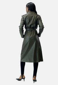 Langer olivgrüner Trenchcoat aus Leder, mit einem taillierten Gürtel, breitem Kragen und glatter Textur, kombiniert mit schlanken schwarzen Hosen und High Heels.