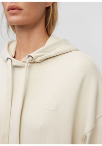Kremowy hoodie z regulowanym sznurkiem, srebrnymi detalami oraz wytłoczonym logo. Miękki materiał w luźnym kroju oraz kieszeń kangurza.