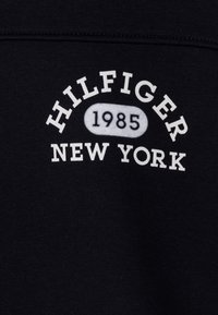 Černá tkanina s bílým logem, které zní "HILFIGER 1985 NEW YORK," s zaobleným textem uspořádaným do oblouku nad hladkým oválem.