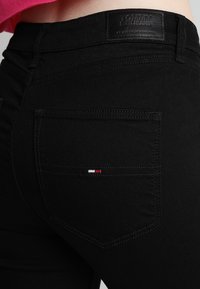 Los jeans de mezclilla negros tienen un bolsillo trasero con un pequeño logotipo rojo, blanco y azul. La cinturilla tiene una etiqueta de piel negra que dice "TOMMY JEANS".