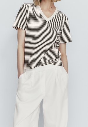 Personne portant un t-shirt à manches courtes en V rayé noir et blanc, rentré dans un pantalon blanc taille haute à jambes larges, les mains dans les poches.