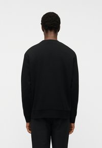 Άνδρας με κοντά κατσαρά μαλλιά, φορούσε μαύρο απλό φούτερ με μακριά μανίκια και μαύρο παντελόνι, στέκεται γυρισμένος με την πλάτη προς τα έξω μπροστά σε λευκό φόντο.