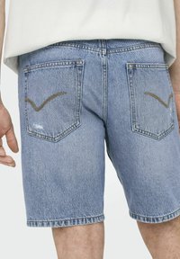 Shorts de mezclilla en azul claro con un dobladillo deshilachado, que presentan dos bolsillos traseros con un patrón de costura curvada en marrón. Llevados con una blusa de color claro.
