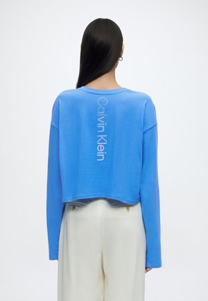 Személy hosszú sötét hajjal, aki kék hosszú ujjú rövid felsőt visel, amelyen fehér, függőleges "Calvin Klein" felirat van a háton, és fehér nadrágot.