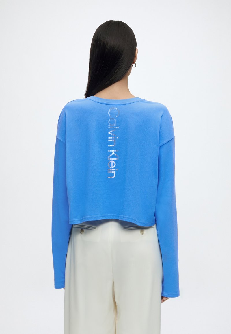 Άτομο με μακριά σκούρα μαλλιά που φοράει μπλε μακρυμάνικη κοντή μπλούζα με κάθετο λευκό κείμενο «Calvin Klein» στην πλάτη και λευκό παντελόνι.