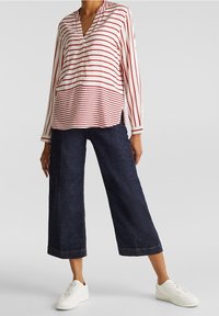 Blouse légère à rayures rouges et blanches avec col en V, manches longues et fentes latérales, associée à un pantalon large foncé et des baskets blanches.