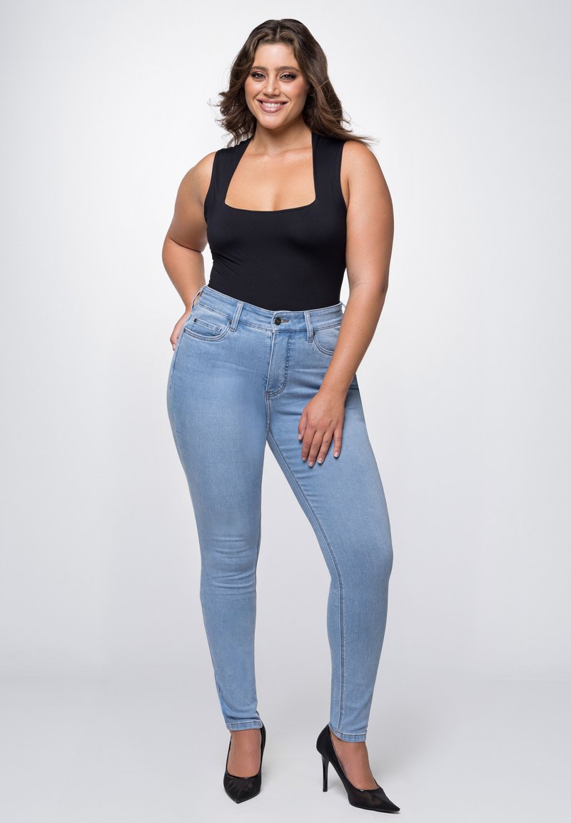 Zwarte bodysuit met vierkante halsuitsnijding, gecombineerd met lichtblauwe skinny jeans. De jeans hebben vijf zakken en een knoopsluiting aan de voorkant, gedragen met zwarte hakken.