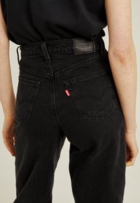 Personne portant un jean Levi’s noir taille haute et une chemise noire, vue de dos, mains détendues le long du corps.