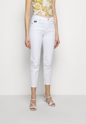 Frau trägt weiße, eng anliegende, gekürzte Jeans, weiße, Riemchensandalen mit hohen Absätzen und ein ärmelloses, gelb gemustertes Crop-Top.