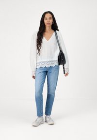 Witte blouse met lange mouwen en kantdetails aan de zoom en manchetten, gecombineerd met lichtblauwe jeans en grijze sneakers, met een zwarte schoudertas.