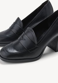 Mocassins en cuir noir avec un bout carré et un talon bloc angulaire. Présente une sangle horizontale avec un accent triangulaire et de fines coutures.