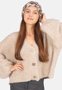 Gilet en tricot beige avec des manches surdimensionnées et trois boutons décoratifs en or. Porté avec un bonnet en imprimé léopard beige. Tissu doux et texturé.