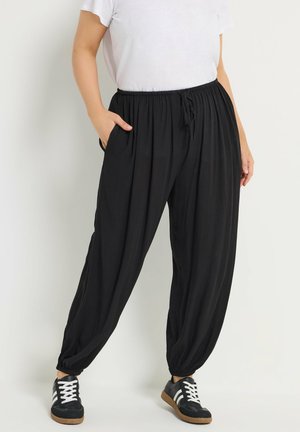 Persona con pantalones jogger negros holgados con puños elásticos, camiseta blanca y zapatillas negras con rayas blancas, de pie frente a un fondo liso.