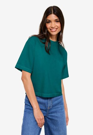 Ung kvinde med langt mørkt hår smiler og bærer en løs, mørkegrøn t-shirt med korte ærmer og blå jeans mod en hvid baggrund.