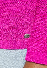 Maglione rosa lavorato a maglia con un motivo testurizzato, dotato di orlo a coste e di un'etichetta in metallo sul lato. Stile casual, materiale leggero.