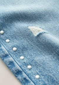 Tissu en denim bleu clair avec une petite déchirure usée et une rangée de clous métalliques ronds blancs le long du bord.