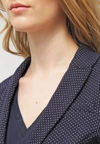 Marinblå kavaj med små vita prickar, slagkrage och en v-ringad topp. Tyget verkar ha en textur, vilket framhäver den sofistikerade designen.