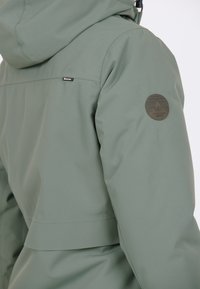 Grön vattentät jacka med huva, en elegant design, en cirkulär badge på ärmen och slät, texturerad väv.