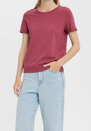 Kobieta nosząca ciemnoróżowy t-shirt z krótkim rękawem i okrągłym dekoltem, noszony na wierzchu jasnoniebieskich, wysokich dżinsów, z jedną ręką w kieszeni.