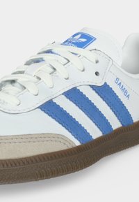 Λευκό αθλητικό παπούτσι Adidas Samba με μπλε ρίγες, λαστιχένια σόλα, λευκά κορδόνια και μπλε λογότυπο Adidas στη γλώσσα και στο πλευρό.