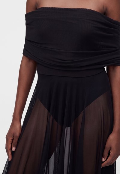 Femme portant un haut noir épaules dénudées et une jupe noire transparente avec des couches inférieures opaques noires, visible de la poitrine aux mains.