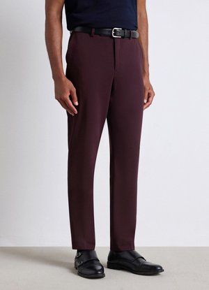 Man wearing dark burgundy tailored trousers, black leather belt, and black leather dress shoes standing on light floor.
