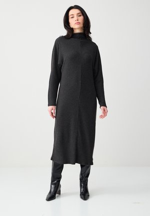 Kobieta stoi, ubrana w długą, ciemnoszarą dzianinową sukienkę z długimi rękawami oraz czarne kozaki za kolano na jednolitym jasnym tle.