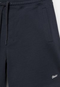 Pantalon de jogging bleu marine foncé avec des poches latérales avant, une taille à cordon de serrage et un petit logo brodé blanc près de l'ourlet.