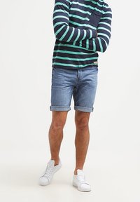 Laivastonsininen ja turkoosin raidallinen pitkähiha paita yhdistettynä vaaleansinisiin farkkushortseihin, käännetyt lahkeet ja valkoiset lenkkarit. Rentoa, väljää istuvuutta.