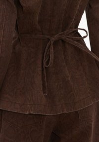 Veste en velours côtelé marron ceinturée à la taille avec une fine ceinture, associée à un pantalon en velours côtelé marron assorti.