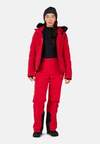 Veste isolée rouge avec une capuche doublée de polaire noire, assortie à un pantalon rouge avec des accents noirs aux genoux. Un t-shirt noir et des bottes complètent l'ensemble.