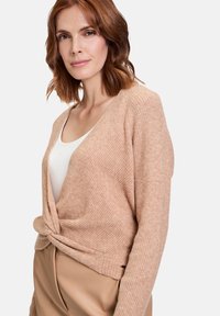 Hellbrauner Strickcardigan mit V-Ausschnitt, Drehdetail an der Taille und langen Ärmeln. Über einem weißen Oberteil mit strukturiertem Finish getragen.