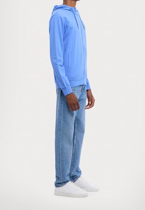 Man draagt een felblauwe hoodie met rits, lichtblauwe spijkerbroek en witte sneakers, staand in profiel tegen een witte achtergrond.