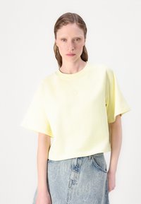 Ανοιχτό κίτρινο, κοντό sweatshirt με ραφές raglan και στρογγυλή λαιμόκοψη. Λείο, μαλακό ύφασμα με μια διακριτική ανάγλυφη λογότυπο. Από κάτω, τζιν φούστα.