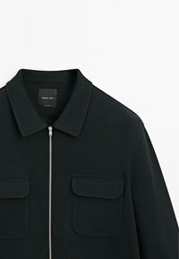 Veste noire avec col, fermeture éclair sur le devant et deux poches poitrines. La texture semble lisse, avec un subtil motif en chevrons.