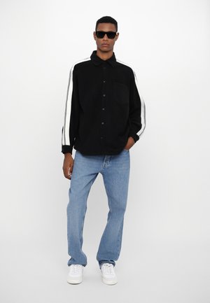 Sort langærmet skjorte med hvide striber, knaplukning, enkelt brystlomme; parret med lyseblå jeans og hvide sneakers.