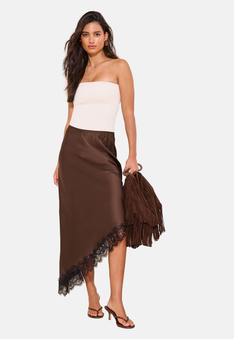 Woman wearing a white strapless top and asymmetrical brown skirt with black lace, holding a brown fringed bag, and wearing brown heeled sandals.