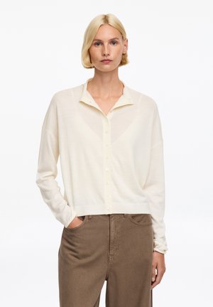 Femme blonde portant un cardigan crème boutonné et un pantalon marron taille haute, debout avec une main dans la poche sur fond blanc.