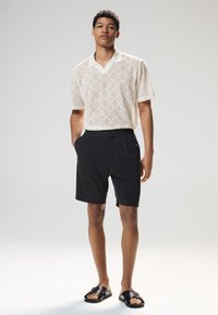 Polo à manches courtes en maille beige clair avec un motif en losange, associé à un short noir et des sandales noires. Texture lisse, coupe décontractée.