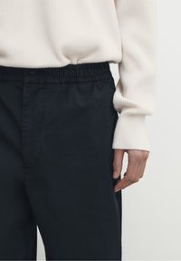 Svarta byxor med elastisk midja gjorda av ett slätt tyg, parat med en ljus beige ribbad tröja; närbild som visar hand och texturdetaljer.