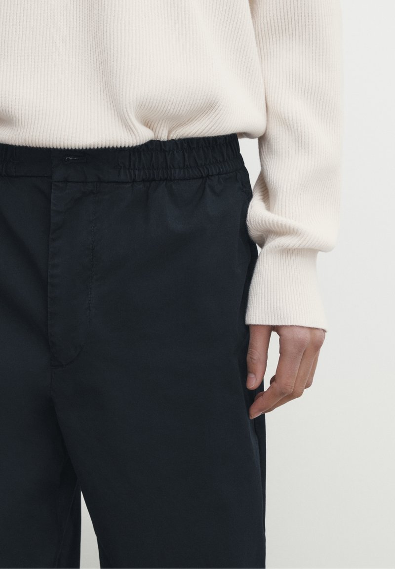 Svarta byxor med elastisk midja gjorda av ett slätt tyg, parat med en ljus beige ribbad tröja; närbild som visar hand och texturdetaljer.