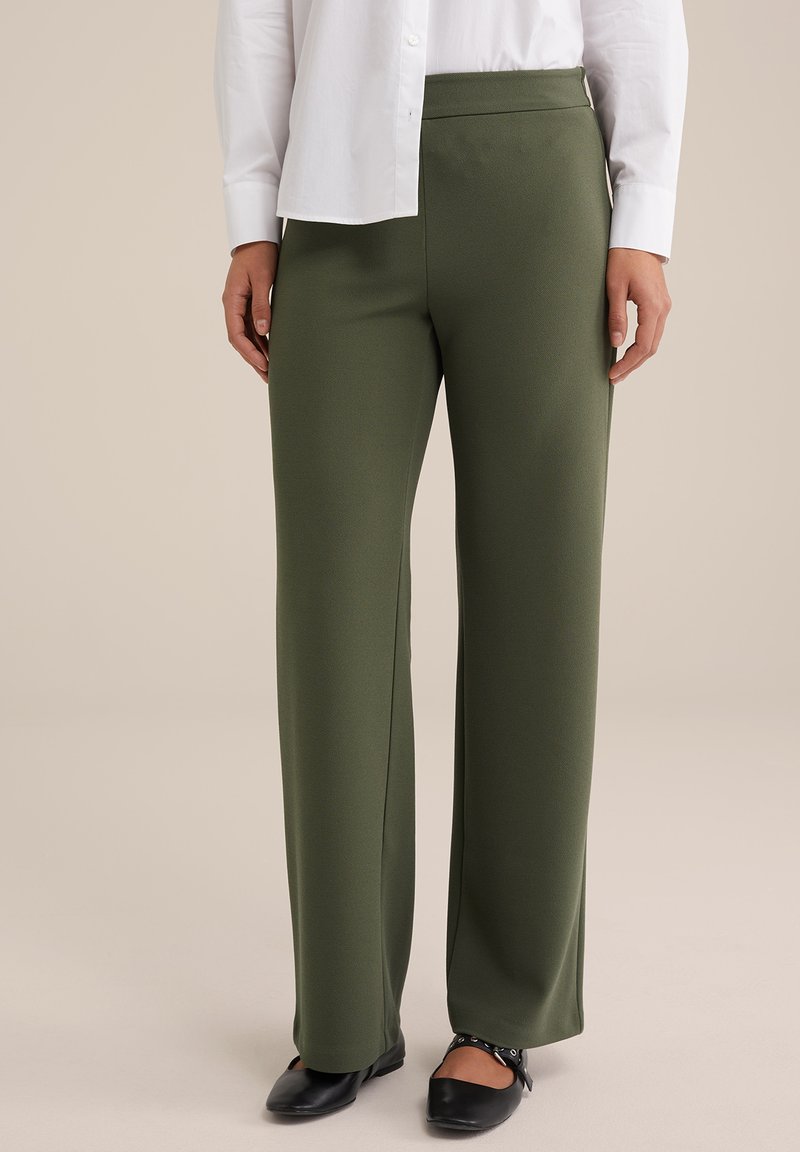 Pantaloni a gamba larga verde oliva in un tessuto morbido e elasticizzato; presentano una vita alta e cuciture ordinate. Abbinati a scarpe nere basse.