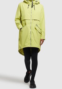 Ljusgrön regnjacka med huva, dragkedja framtill, stora fickor, indragen midja och längre bakre fåll, tillverkad av vattentätt material.