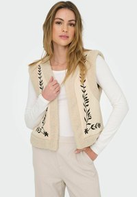 Krämfärgad väst med fleecestuktur, svart blommönster broderat på varje sida, bärs över en vit långärmad topp, i kombination med ljusa byxor.