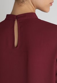 Bordeauxblus med hög krage, nyckelhålsöppning bak, mjukt tyg och rena sömmar. Tyget faller fluid över axlarna.